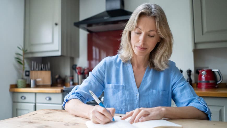 Tagebuch schreiben ist beliebter denn je. Frau sitzt am Küchentisch und schreibt in einem Buch.