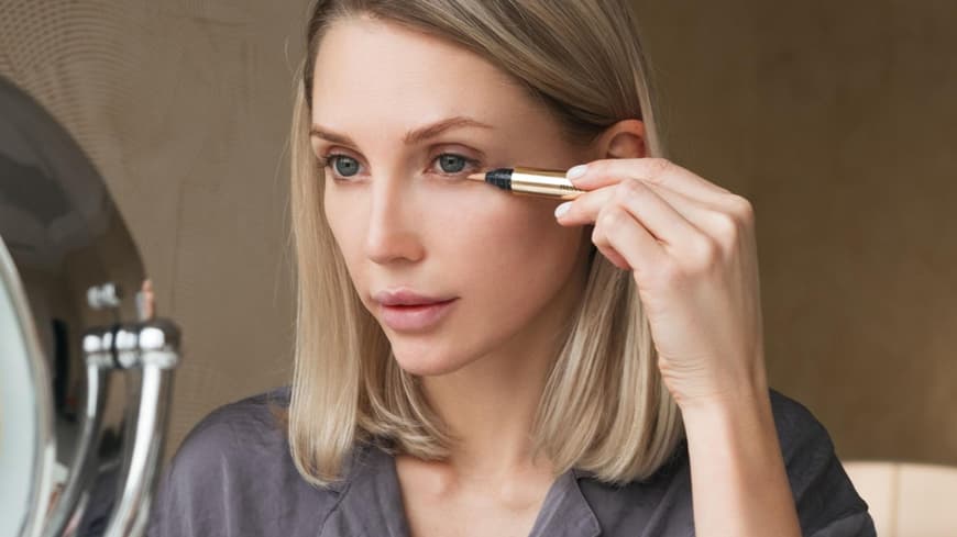 Ein guter Concealer sollte sich nicht absetzen. Frau sitzt vor dem Spiegel und trägt Concealer auf.