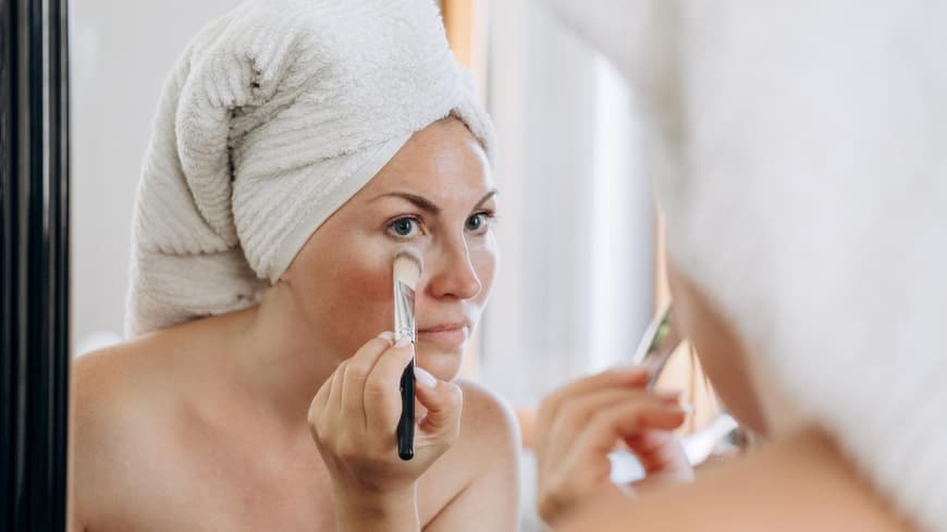 Augenringe sind bei vielen Frauen ein leidiges Thema. Frau schminkt sich mit einem Pinsel und deckt Augenringe ab.