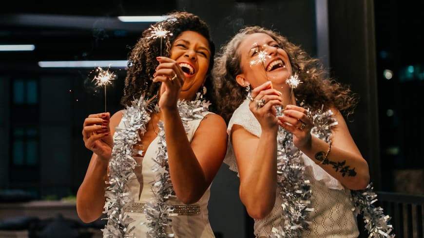 Silvester verbringen viele mit Freunden. Zwei Frauen mit Wunderkerzen und Glitzerdeko um den Hals lachen.