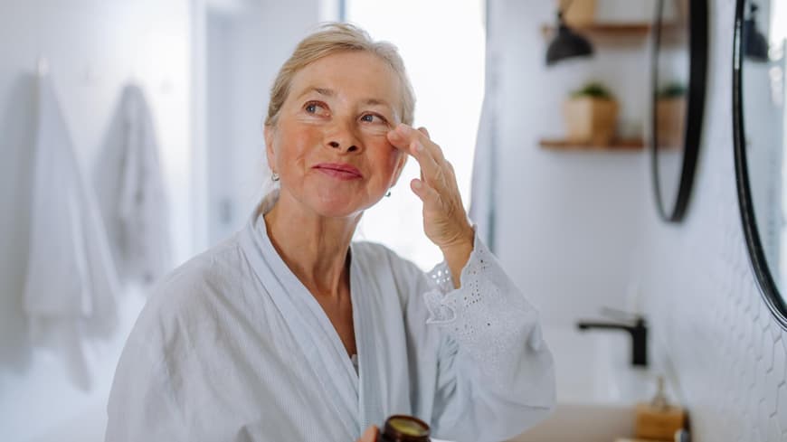 Die beste Augencreme ab 50 kann helfen, Falten zu reduzieren. Ältere Frau trägt im Bademantel eine Creme unter ihren Augen auf.