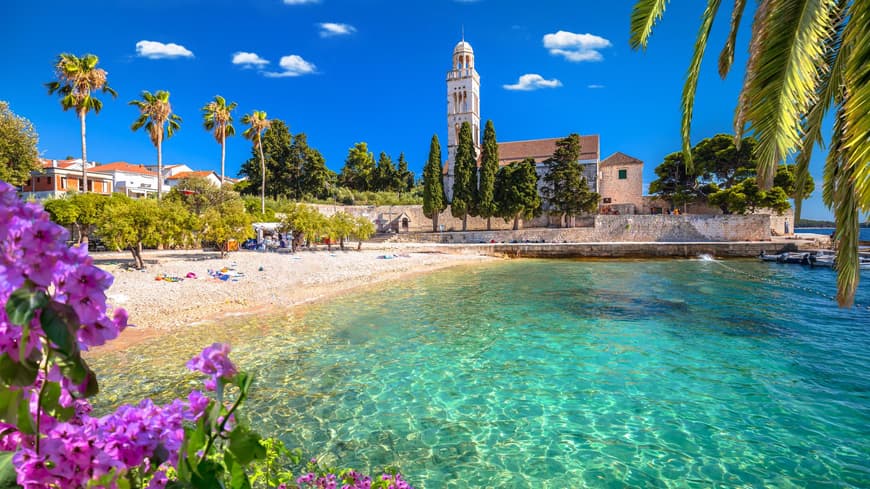 Die Insel Hvar hat viele schöne Strände zu bieten. Strand mit ein paar Handtüchern, Palmen und einer Kirche im Hintergrund