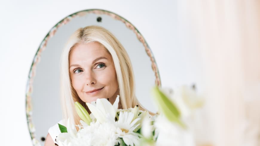 Wie du einen Grünstich mit Hausmitteln entfernen kannst, erfährst du hier. Blonde Frau schaut in den Spiegel