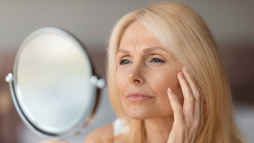 Blotting Sticks können bei glänzender Haut Wunder bewirken. Ältere Frau schaut skeptisch in einen Handspiegel und fässt sich mit der Hand ins Gesicht.