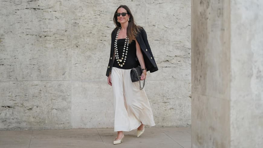 Maxi-Röcke im Boho-Stil liegen wieder total im Trend. Frau trägt einen weißen Maxi-Rock im Boho-Stil mit einem schwarzen Top und Blazer und zwei großen Perlenketten.