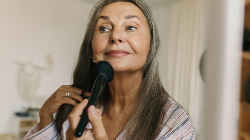 Beim Sommer-Make-up können uns einige Schminkfehler schnell älter aussehen lassen. Ältere Frau mit grauen Haaren sitzt vor einem Spiegel und geht mit einem Puderpinsel über ihr Kinn.