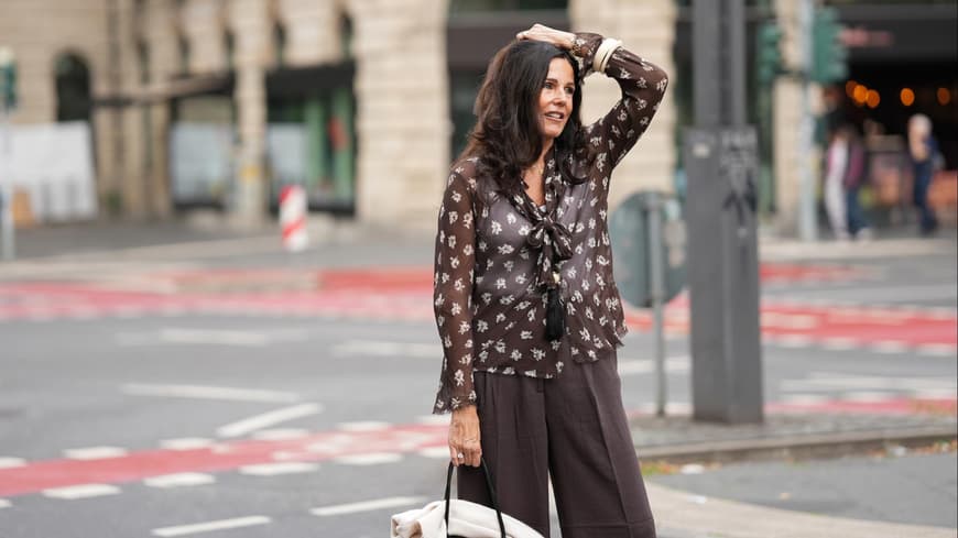 Dunkelbraune Hosen liegen im Trend. Frau mit dunkelbraunen Haaren trägt eine dunkelbraune Hose und transparente Bluse mit Muster und fässt sich mit einem Arm an den Kopf..