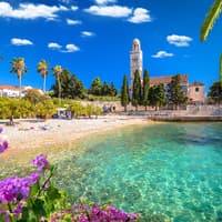 Die Insel Hvar hat viele schöne Strände zu bieten. Strand mit ein paar Handtüchern, Palmen und einer Kirche im Hintergrund