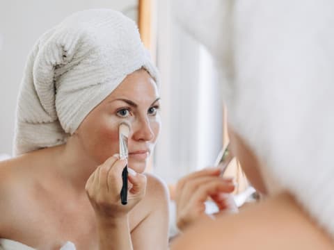Augenringe sind bei vielen Frauen ein leidiges Thema. Frau schminkt sich mit einem Pinsel und deckt Augenringe ab.