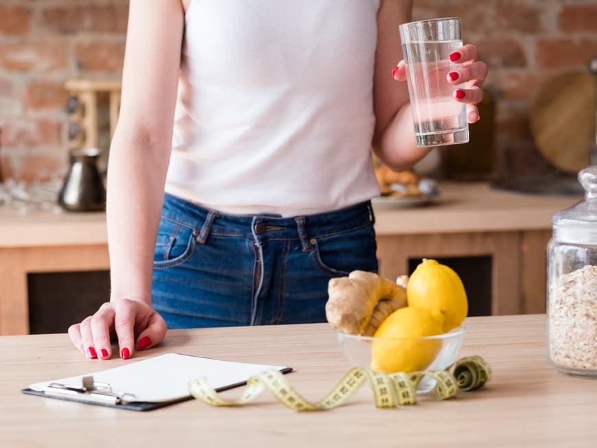Abnehmen muss kein Hexenwerk sein. Frau hält ein Glas Wasser in der Hand und steht am Tisch mit einem Maßband, Zettel, Ingwer und Zitrone.