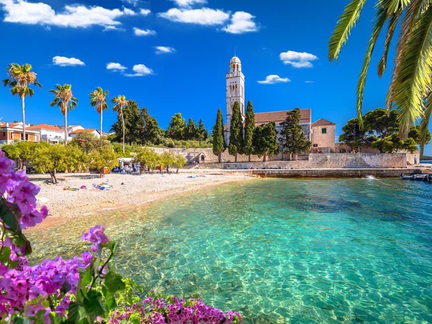 Die Insel Hvar hat viele schöne Strände zu bieten. Strand mit ein paar Handtüchern, Palmen und einer Kirche im Hintergrund