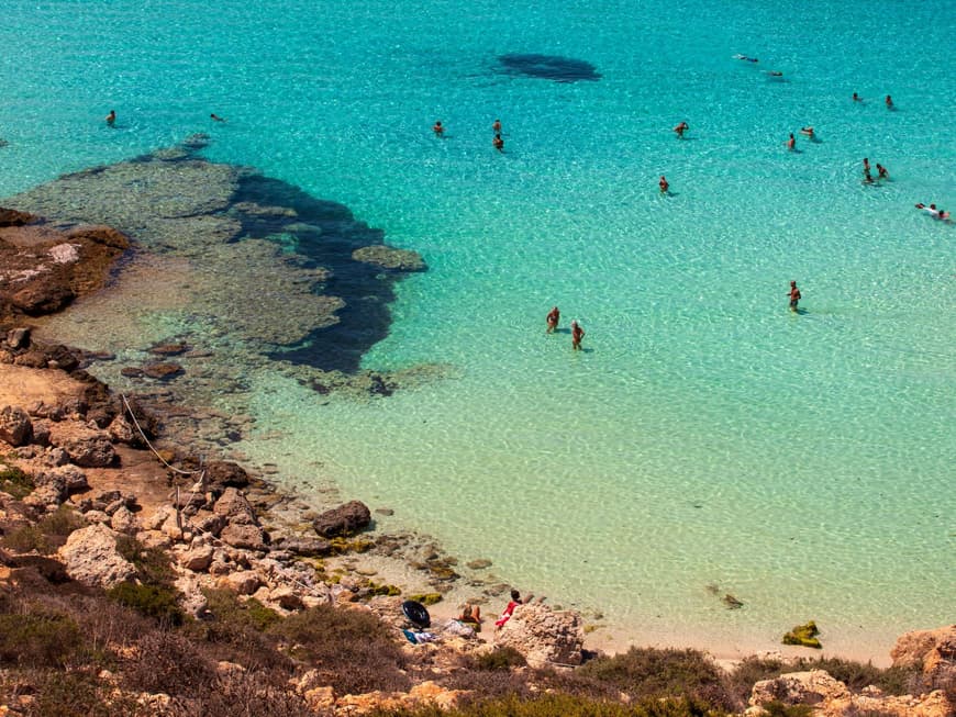 Das Wasser am Kaninchenstrand auf Lampedusa ist Türkisblau. Ein Strand mit Felsen und Menschen im Wasser.